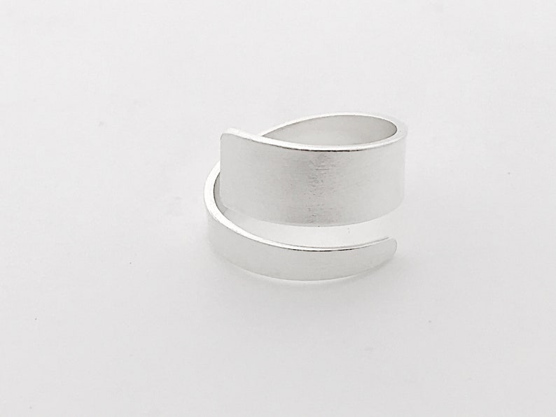 May include: A silver ring with a wide, flat band that wraps around the finger. The band is smooth and has a brushed finish.