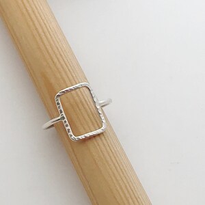 May include: A silver ring with a textured, rectangular shape. The ring is open on one side and closed on the other.