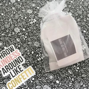 May include: A white organza bag containing a white card. The card has the text "JEWELLERY BY DAPRI" printed on it. The bag is placed on a black and white floral patterned surface. A sticker with the text "THROW KINDNESS AROUND LIKE CONFETTI" is also on the surface.