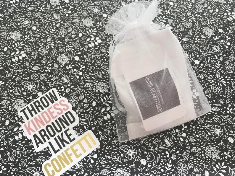 May include: A white organza bag containing a white card. The card has the text "JEWELLERY BY DAPRI" printed on it. The bag is placed on a black and white floral patterned surface. A sticker with the text "THROW KINDNESS AROUND LIKE CONFETTI" is also on the surface.