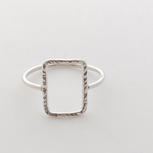 May include: A silver ring featuring a textured, rectangular shape. The ring is open on one side, creating a frame-like design.