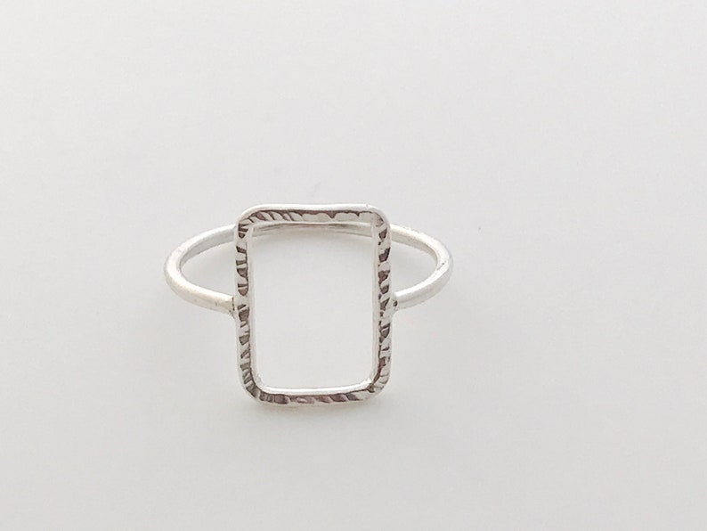May include: A silver ring featuring a textured, rectangular shape. The ring is open on one side, creating a frame-like design.