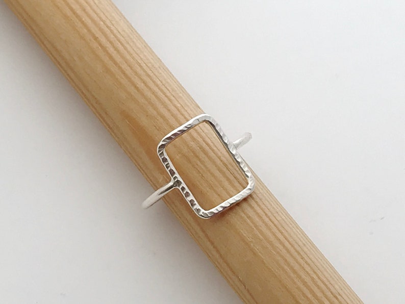 May include: A silver ring with a textured, rectangular shape. The ring is open on one side and is designed to be worn on a finger.