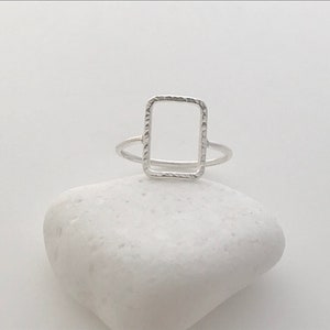 May include: A silver ring with a textured, rectangular frame design.