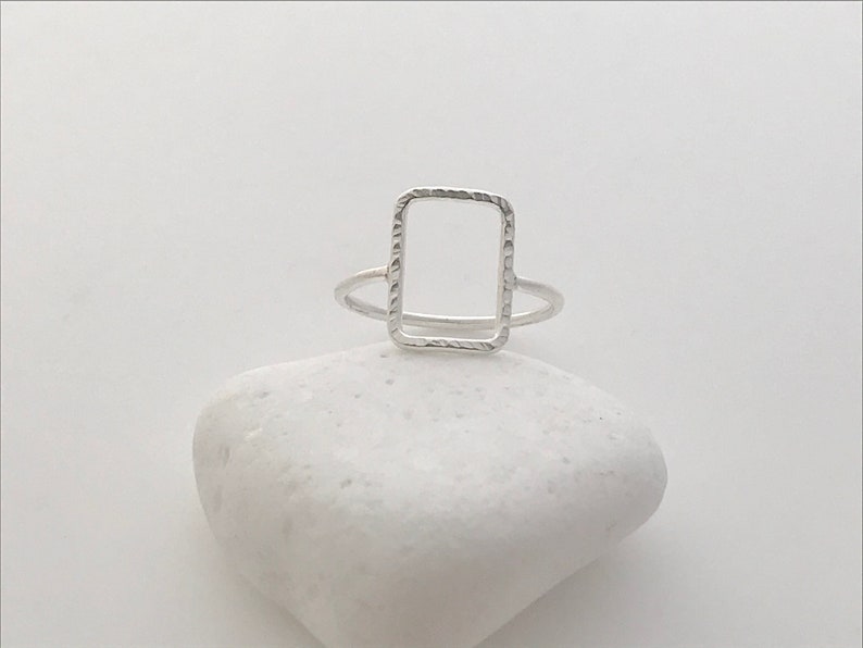 May include: A silver ring with a textured, rectangular frame design.