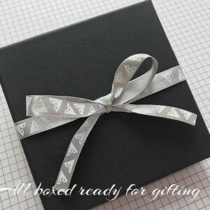 May include: A black gift box with a silver ribbon tied in a bow. The ribbon has a white geometric pattern. The text "All boxed ready for gifting" is written on the bottom of the image.