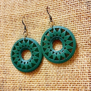 May include: A pair of green earrings with a geometric design. The earrings are made of wood and have a circular shape with a hole in the centre. The earrings are hanging from a brown metal hook.