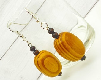 May include: A pair of wooden earrings with a natural wood grain finish. The earrings feature large, oval-shaped beads with horizontal stripes, connected by silver-colored hooks and small, dark brown beads. The earrings are displayed against a blurred background.