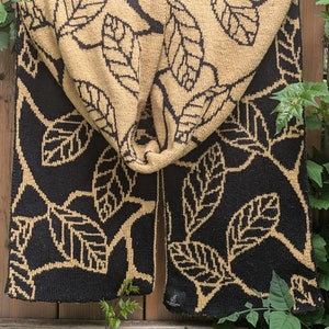 Peut inclure: Écharpe tricotée noire et beige avec un motif de feuilles. L'écharpe est drapée sur une clôture en bois. Le motif de feuilles est beige sur fond noir. L'écharpe est faite d'une matière douce et texturée.