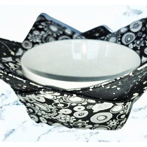 May include: A white ceramic bowl rests inside a black and white patterned fabric bowl cozy. The cozy has a floral and geometric design. The bowl and cozy are set against a white background.