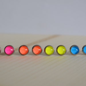 May include: A set of eight small, round earrings with neon colored glass inserts. The earrings are arranged in a line on a wooden surface. The colors are pink, orange, yellow, and blue.