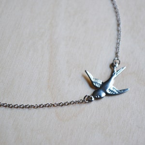 May include: A silver chain necklace with a silver bird pendant. The bird is in flight with its wings spread out.