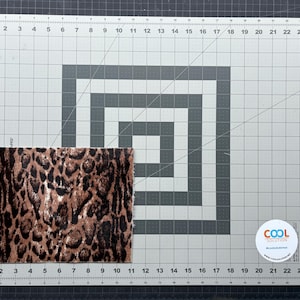 May include: A rectangular piece of fabric with a brown and black leopard print pattern. The fabric is placed on a gray cutting mat with grid lines and measurements in inches.