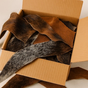 May include: A cardboard box filled with various pieces of cowhide leather. The leather pieces display a range of colors, including brown, black, and gray, with natural hair patterns. The box is open, revealing the contents.