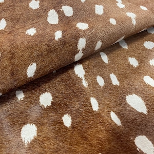 May include: A close-up of a brown hide featuring a pattern of white spots, reminiscent of a deer's coat. The material's texture is evident, with the spots differing in size and form. The hide is folded, exposing the reverse side.