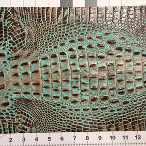 May include: Close-up of a teal and brown faux crocodile skin fabric. The fabric has a textured, embossed pattern that resembles crocodile scales.