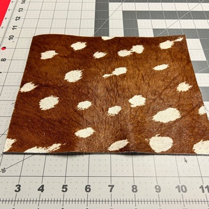 May include: A rectangular piece of brown cowhide with a pattern of white spots. The hide is on a gray and white grid surface, with a red edge visible in the upper left corner. The hide is approximately 25 cm by 30 cm.