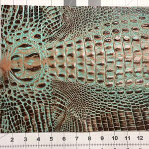 May include: A close-up of a turquoise and brown faux crocodile skin fabric. The fabric has a textured, embossed pattern that resembles crocodile scales.