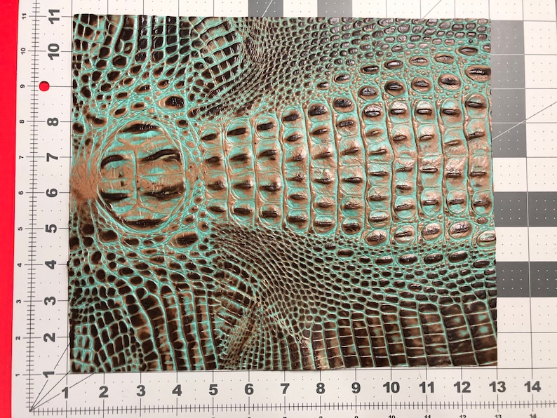 May include: A close-up of a turquoise and brown faux crocodile skin fabric. The fabric has a textured, embossed pattern that resembles crocodile scales.