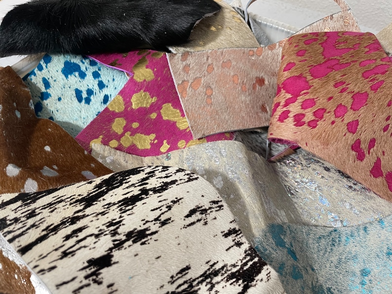 May include: A collection of cowhide rugs in various colors and patterns, including black, brown, white, pink, blue, and gold. The rugs are arranged in a pile, showing off their unique textures and designs.