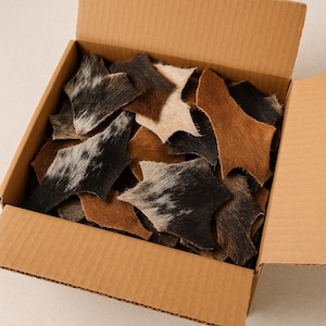 May include: A cardboard box filled with irregularly shaped pieces of animal hide. The hides are a mix of colors, including black, white, brown, and tan. The pieces have a soft, textured appearance.