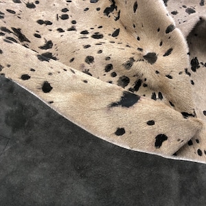 Puede incluir: Primer plano de una alfombra de piel de vaca con manchas blancas y negras. La alfombra est&aacute; doblada por la mitad, revelando un respaldo de ante marr&oacute;n oscuro.