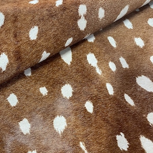 May include: A close-up of a brown and white spotted hide. The hide has a natural texture with a pattern of irregular white spots against a brown background. The material is folded, showing the texture and depth of the material.