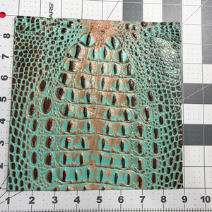 May include: A close-up of a turquoise and brown faux crocodile skin fabric. The fabric has a textured, embossed pattern that resembles crocodile scales.