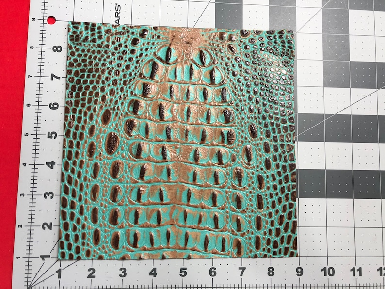 May include: A close-up of a turquoise and brown faux crocodile skin fabric. The fabric has a textured, embossed pattern that resembles crocodile scales.