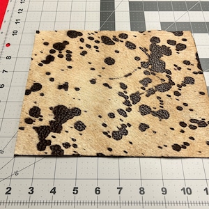 Puede incluir: Primer plano de un trozo de piel de vaca marr&oacute;n y blanca con manchas negras. La piel de vaca est&aacute; sobre una superficie cuadriculada gris y blanca.