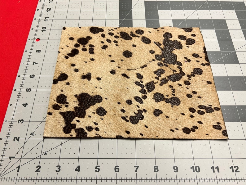 Puede incluir: Primer plano de un trozo de piel de vaca marr&oacute;n y blanca con manchas negras. La piel de vaca est&aacute; sobre una superficie cuadriculada gris y blanca.