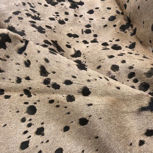 Puede incluir: Primer plano de una alfombra de piel de vaca con fondo marr&oacute;n claro y manchas negras. La alfombra tiene una superficie suave y texturizada.