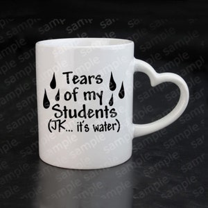Puede incluir: Taza de cerámica blanca con asa en forma de corazón. La taza tiene texto negro que dice "Tears of my Students (JK... it's water)" y cuatro gotas de lluvia negras.
