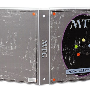 May include: A white binder with a grey and black cover. The cover has the text "MTG DECK COLLECTION" and a black circle with the text "MTG" in white. The binder is closed.