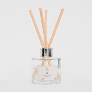 May include: A clear glass diffuser bottle with a silver collar and light brown reeds. The bottle contains a clear liquid and a label that reads "Teakwood & Vanilla". The reeds are arranged in a fan shape, creating a decorative and aromatic home fragrance.