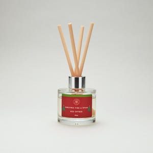May include: A clear glass reed diffuser with a silver cap and wooden reeds. The label reads "Christmas Pine & Spices Reed Diffuser 100ml" in red and white. The diffuser contains a clear liquid.