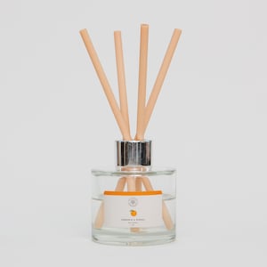 May include: A clear glass diffuser bottle with a silver cap and pale reeds. The bottle features a white label with the words "Mandarin & Mimosa" and a citrus fruit graphic. The reeds are arranged to fan out from the top of the bottle.