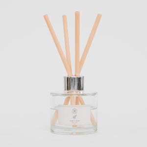 May include: A clear glass essential oil diffuser with a silver collar and several light brown reeds. The diffuser contains a clear liquid and a label with text and a botanical illustration. The reeds are arranged to fan out from the top.