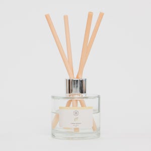 May include: A clear glass diffuser bottle with a silver collar and beige reeds. The bottle contains a clear liquid and a label that reads "Lemon Drizzle". The reeds are arranged to fan out from the top of the bottle.