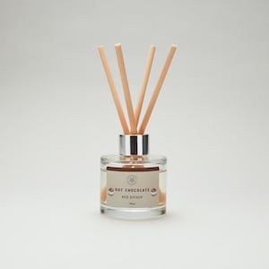 May include: A glass reed diffuser with a silver cap and wooden reeds. The label reads "HOT CHOCOLATE REED DIFFUSER". The bottle contains a light brown liquid. The reeds are arranged in a fan shape. The bottle holds 100ml.