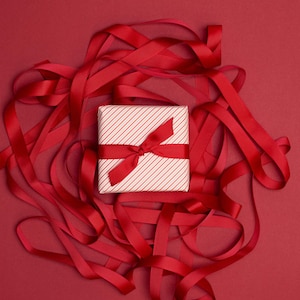 Könnte beinhalten: Eine quadratische Geschenkbox, eingewickelt in weißes Papier mit dünnen diagonalen roten Streifen, gebunden mit einer einfarbigen roten Schleife. Die Box liegt inmitten eines chaotischen Gewirrs aus langen, roten Stoffbändern auf rotem Grund.