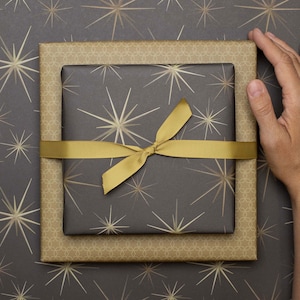 Könnte beinhalten: Zwei quadratische Geschenkboxen, die übereinander gestapelt sind und in dunkelgraues und goldenes Papier mit Sternenmuster eingewickelt wurden. Die obere Box ist mit einem einfarbigen goldenen Band versehen. Das Geschenkpapier hat ein geometrisches Sternmuster.