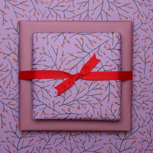 Könnte beinhalten: Zwei quadratische Geschenkboxen, die übereinander gestapelt sind. Die untere Box ist in violettes Papier mit einem feinen Gittermuster eingewickelt. Die obere Box ist in violettes Papier mit einem braunen Zweigmuster und orangen Beeren eingewickelt.