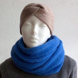 Op de afbeelding: Een mannequinhoofd draagt een lichtbruine tulband-stijl hoed en een felblauwe infinity sjaal. De sjaal is gemaakt van een zacht, gebreid materiaal. De achtergrond is een effen witte muur.