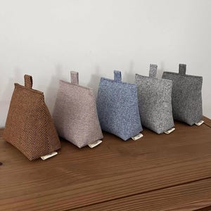 May include: Five fabric doorstops in brown, mauve, blue, grey, and dark grey. Each has a small loop at the top and a rectangular tag at the bottom. They are arranged on a wooden surface.