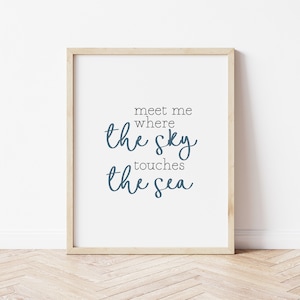 May include: A framed print with the words "meet me where the sky touches the sea" in a blue script font. The print is in a light wood frame and is displayed against a white wall.