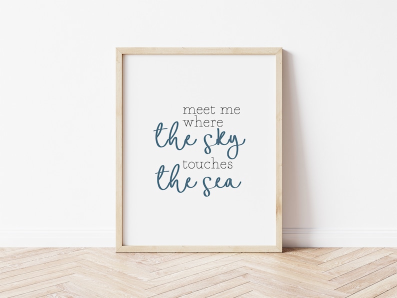 May include: A framed print with the words "meet me where the sky touches the sea" in a blue script font. The print is in a light wood frame and is displayed against a white wall.