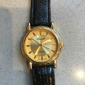 Op de afbeelding: Een gouden polshorloge met een zwarte leren band. De wijzerplaat van het horloge is goudkleurig met zwarte wijzers en zwarte Romeinse cijfers als uurmarkeringen. Op de wijzerplaat van het horloge staan ook de woorden "Rockwell" en "Quartz" gedrukt.