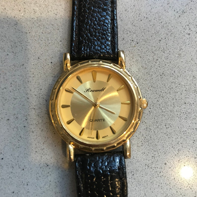 Op de afbeelding: Een gouden polshorloge met een zwarte leren band. De wijzerplaat van het horloge is goudkleurig met zwarte wijzers en zwarte Romeinse cijfers als uurmarkeringen. Op de wijzerplaat van het horloge staan ook de woorden "Rockwell" en "Quartz" gedrukt.