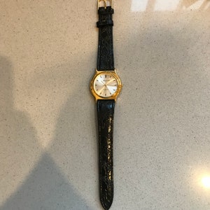 Op de afbeelding: Een gouden polshorloge met een witte wijzerplaat en een zwarte leren band. De wijzerplaat van het horloge heeft zwarte Romeinse cijfers als uurmarkeringen en een datumscherm op 3 uur.